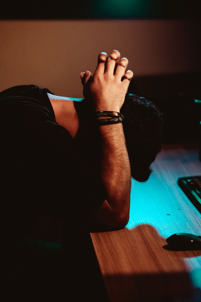 A man slumped at his desk at night, hands clasped over his head