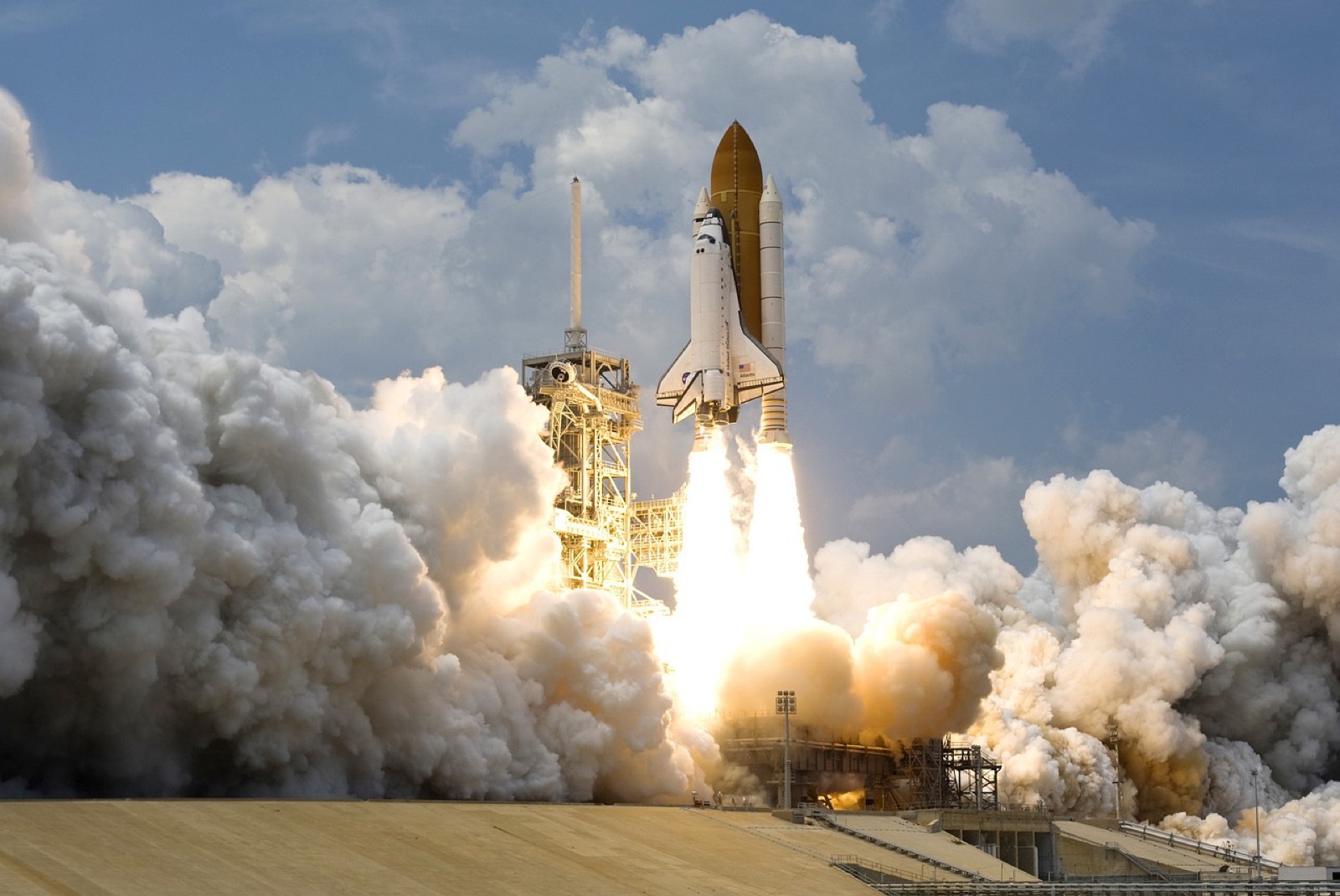 A space shuttle at liftoff, engines at full force, smoke and steam billowing around the launch pad